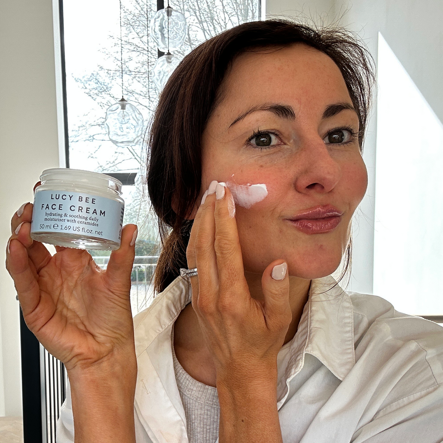 Woman applying face cream from a jar labeled 'Lucy Bee' in a bright room.