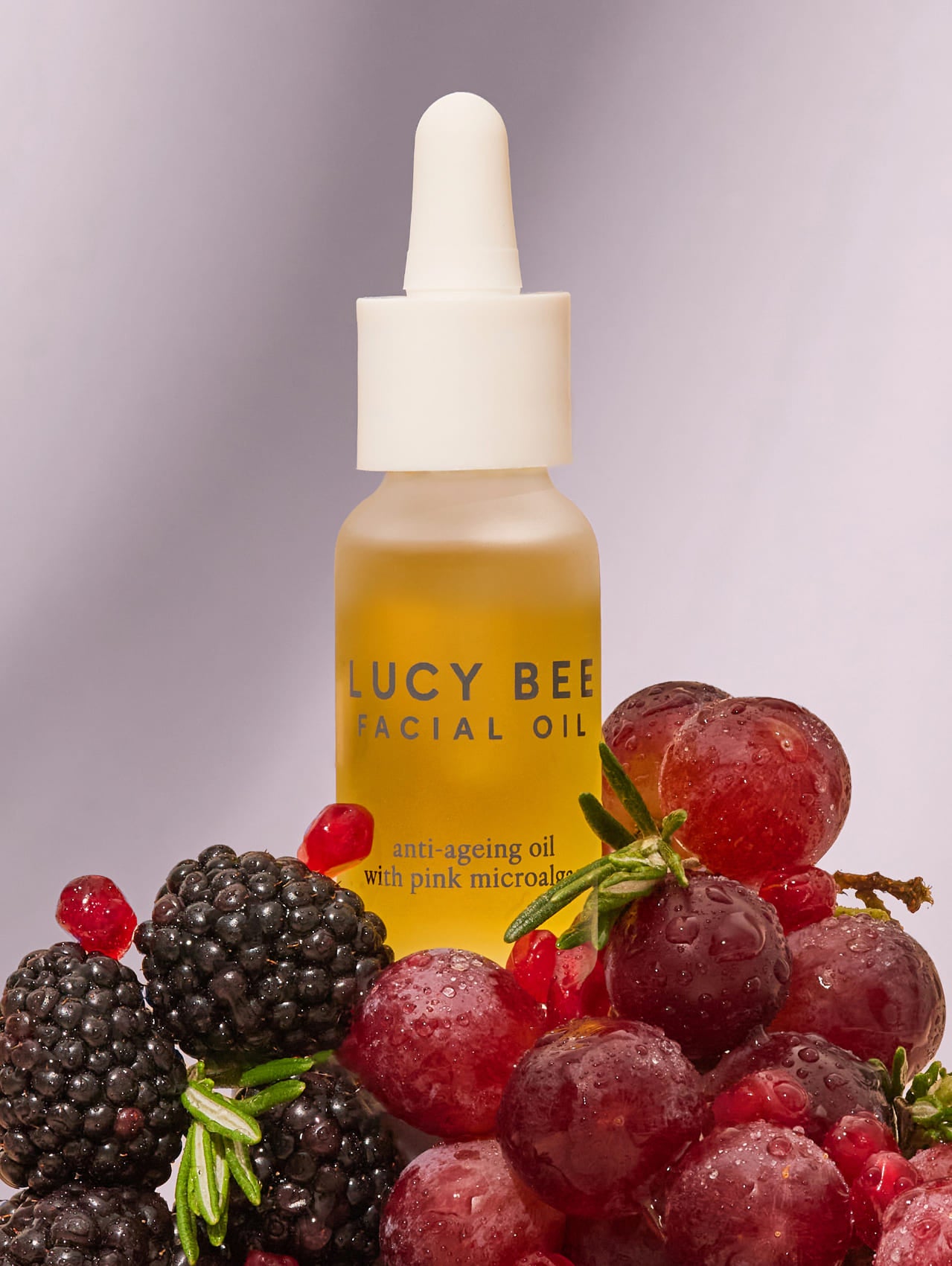 Bottle of Lucy Bee facial oil with a dropper surrounded by grapes and blackberries on a gray background