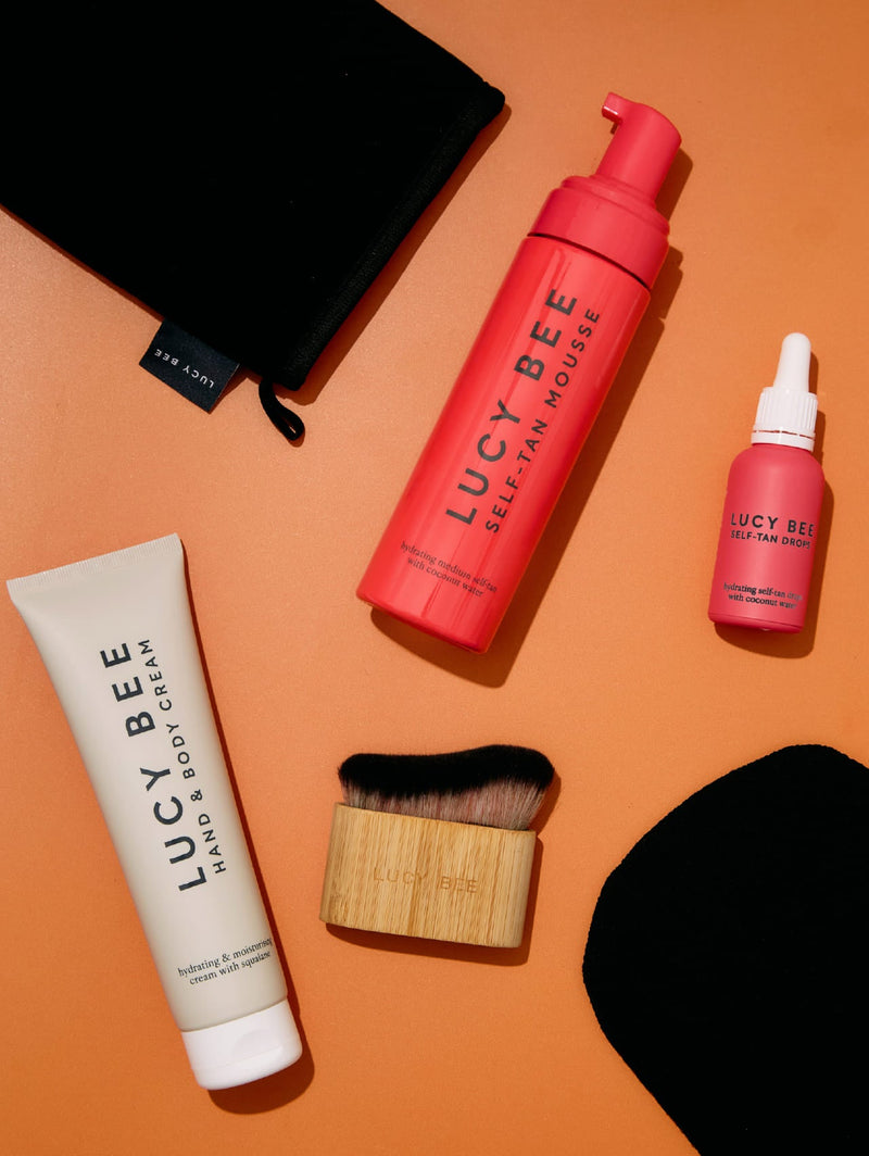 Lucy Bee self-tan mousse, tan drops, hand and body cream, tanning mitt and tan brush laid out on an orange background